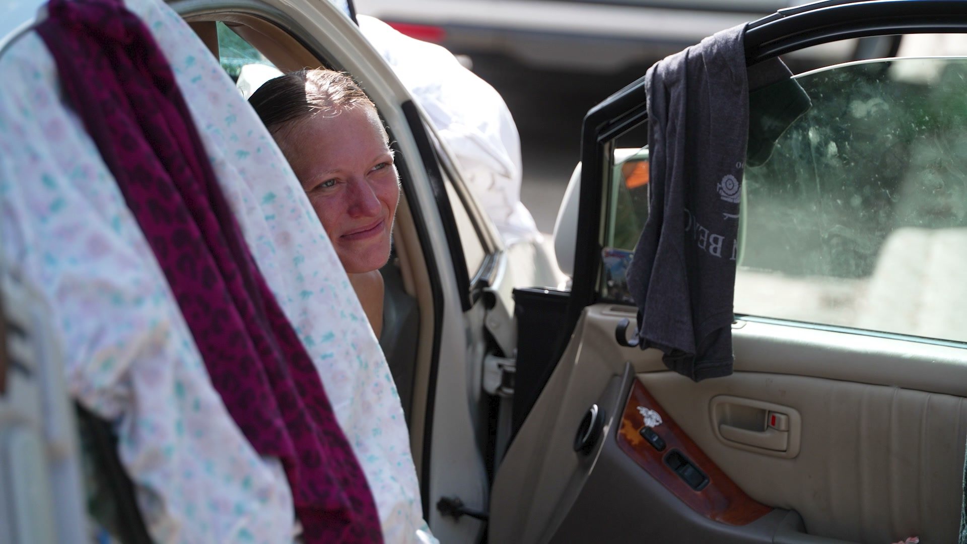 Sarah looks out from behind the open door of the Lexus, laundry hung over the window