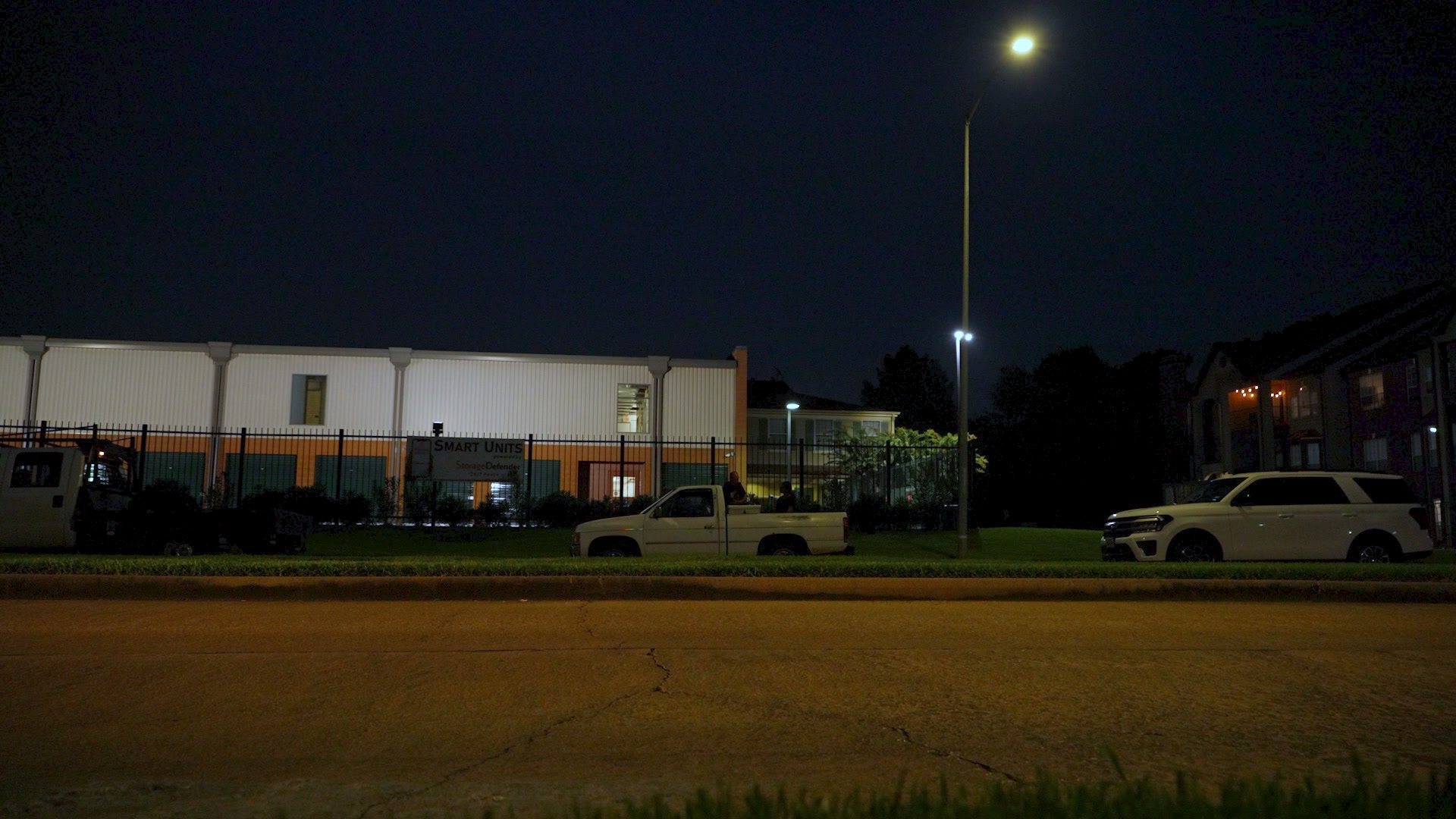 Night. A row of parked vehicles along a fence under streetlights, a small white pickup truck in the middle