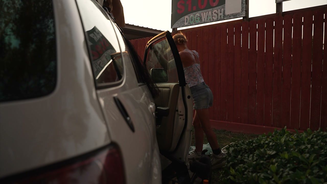 Sarah walks along a red fence beside the open door of the Lexus, a car wash sign above
