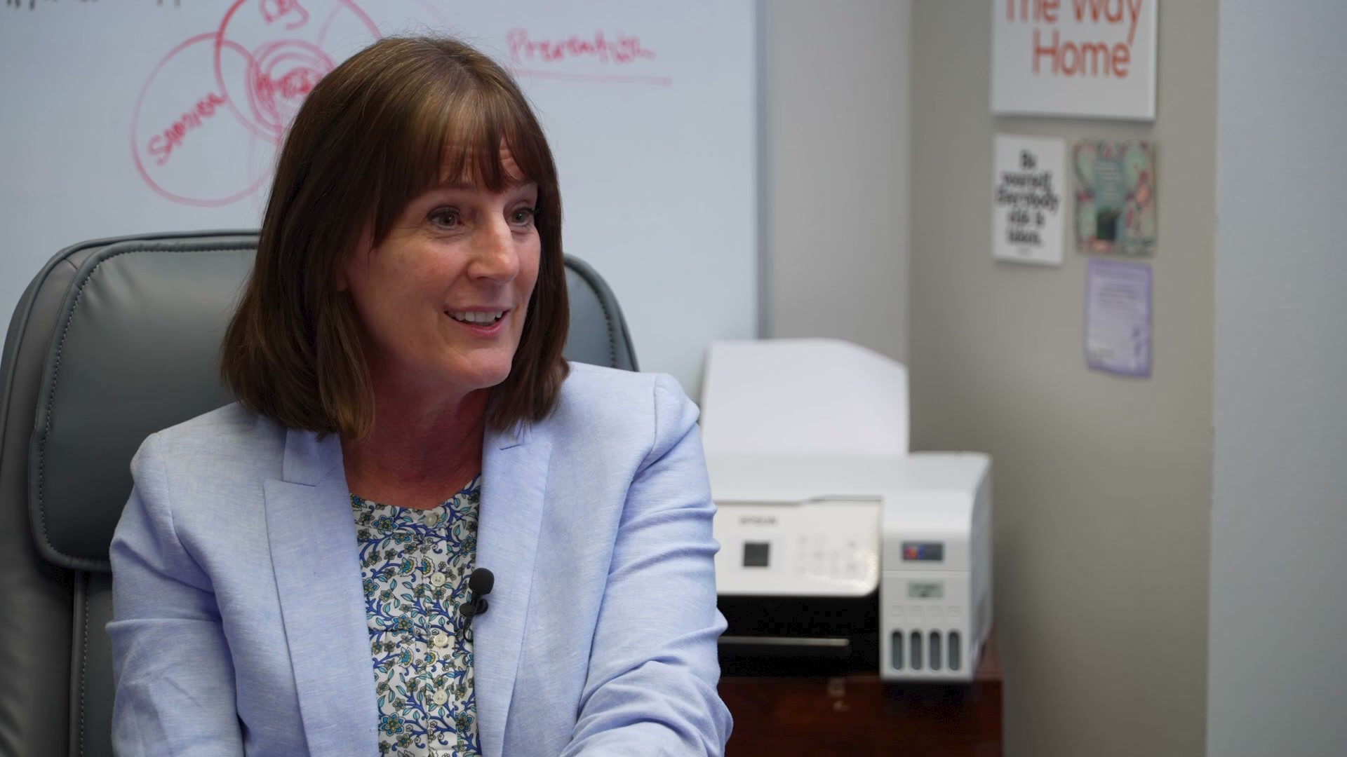 A woman in a light blue blazer smiles during an interview in an office