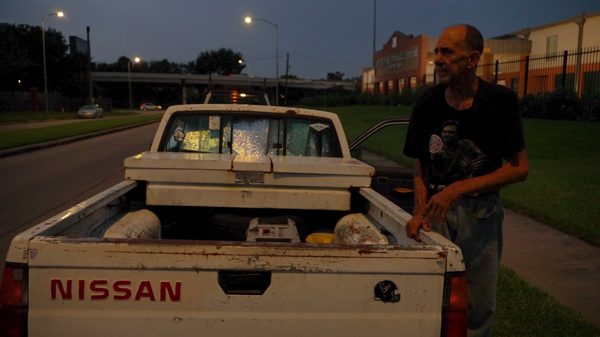 Gary stands at the tailgate of his white Nissan pickup at dusk under streetlights