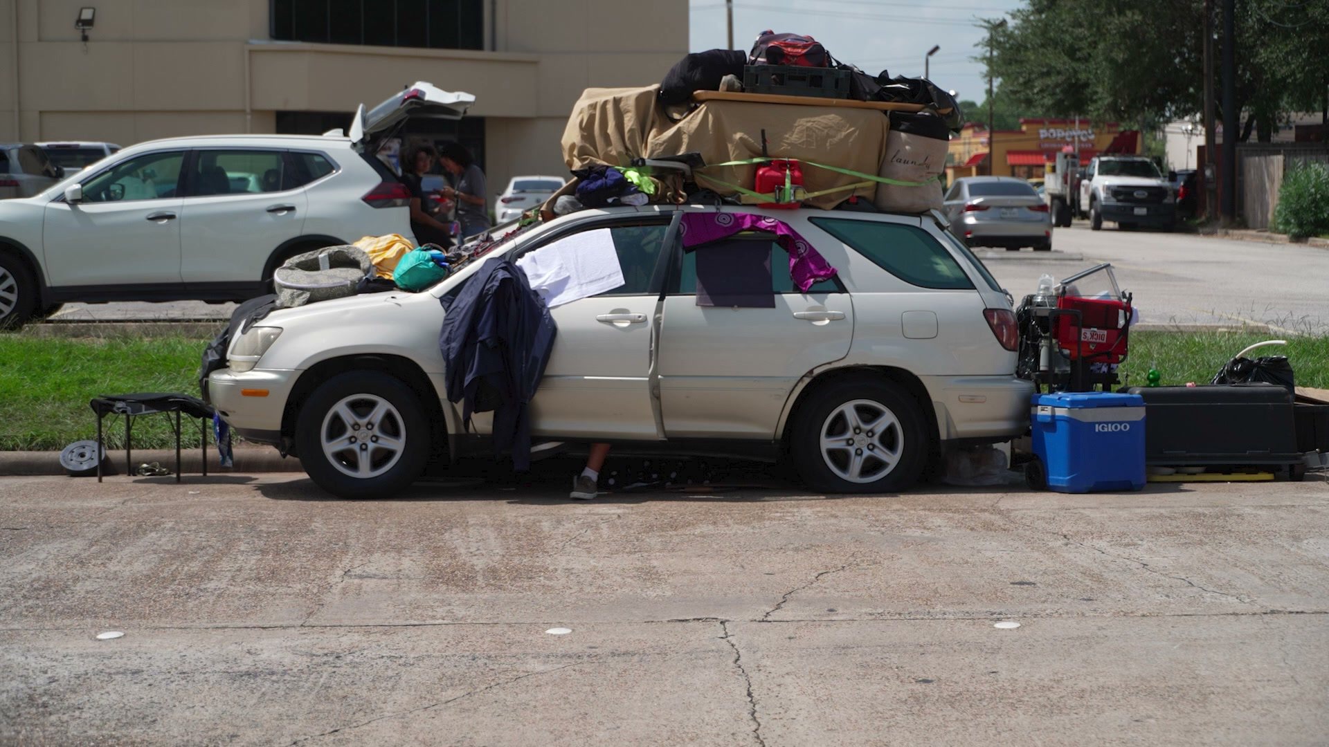 A champagne Lexus SUV parked at a curb, its roof piled with bags and tarps, clothes over the windows, a cooler and gear beside it
